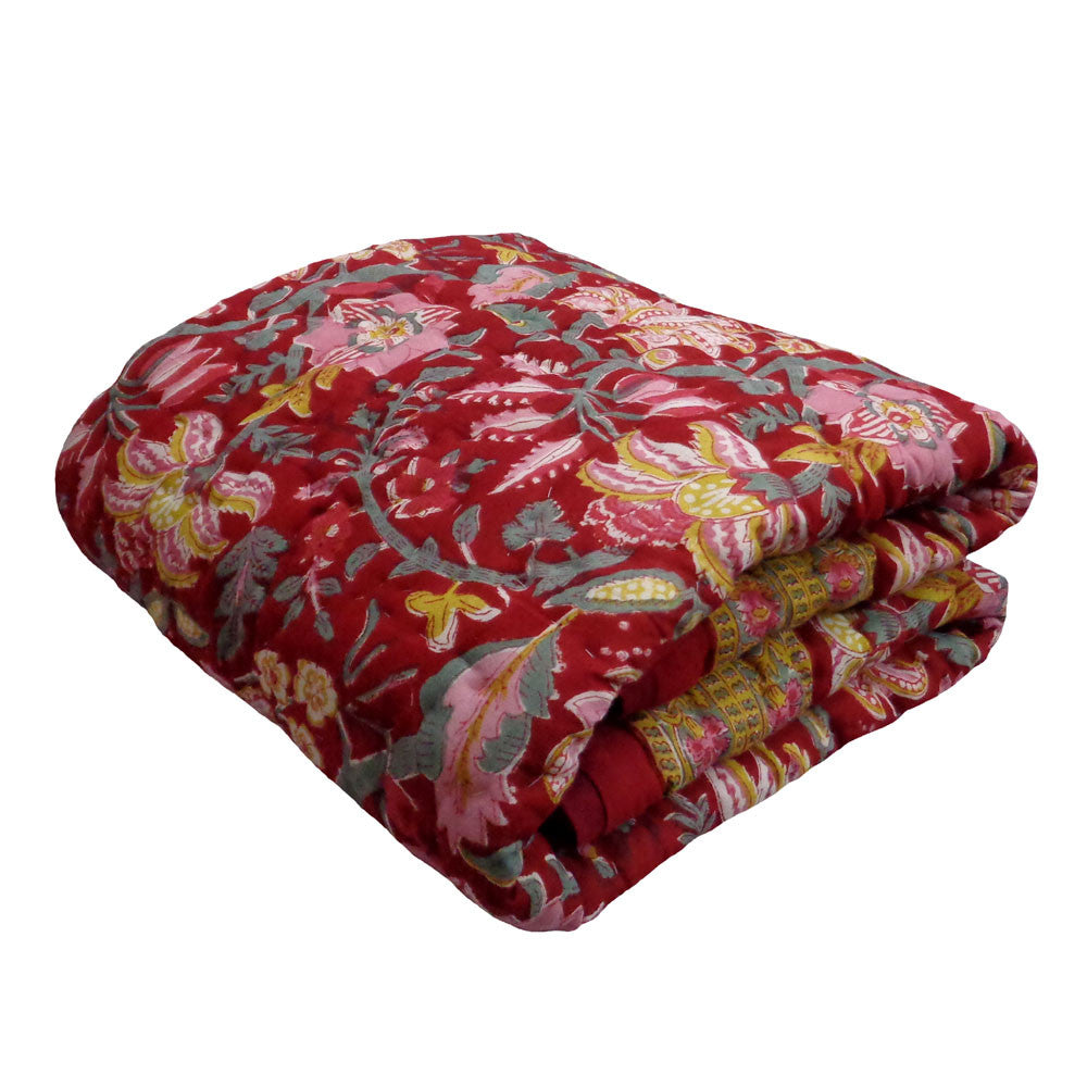 A folded quilted blanket featuring a vibrant red base with a detailed floral and botanical pattern in pink, yellow, and sage green, set against a plain white background.