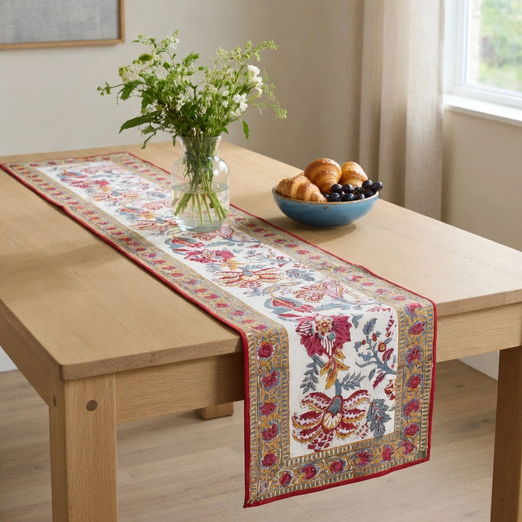 A rectangular table runner with a red border and a detailed floral and botanical pattern in red, blue, green, and gold, displayed on a light wood dining table. The table is styled with a glass vase of white flowers and a blue bowl of pastries.