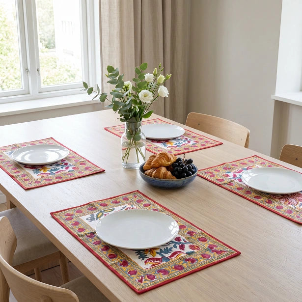 Four rectangular table placemats with a colorful floral and geometric pattern and red borders, arranged on a light wood dining table set with white plates, a vase of flowers, and a bowl of pastries.