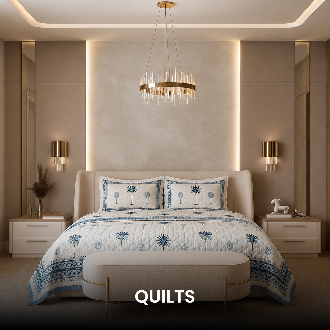A neatly made bed in a bright bedroom setting, covered with a white quilted bedspread featuring a decorative floral border in muted pink and sage green, with matching decorative pillows.