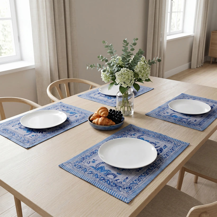 A dining table set with four blue patterned placemats, white plates, a vase of flowers, and a bowl of pastries in a bright, neutral-toned dining room.