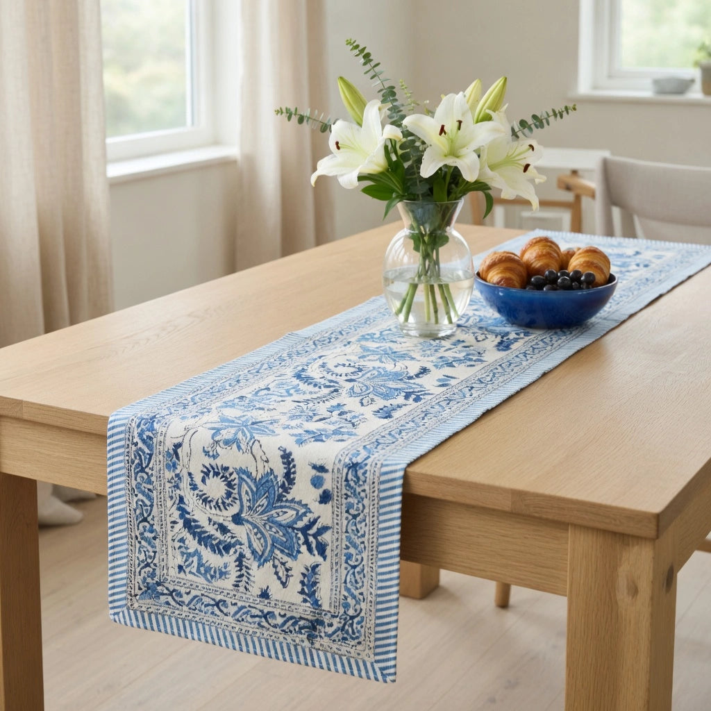 A blue and white patterned table runner draped over a light wood dining table, styled with a glass vase of white lilies and a blue bowl of pastries in a bright, modern room.