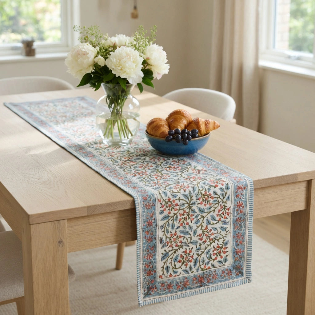 A rectangular table runner with a vintage-style floral pattern in blue, red, and beige, styled on a light wood dining table with a vase of white flowers and a bowl of pastries.
