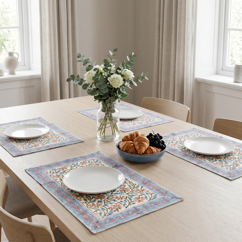 Four rectangular table placemats with a light blue border and intricate multicolor floral patterns are arranged on a light wood dining table. Each placemat holds a white plate, and the table is styled with a vase of white flowers and a bowl of croissants and grapes in a bright, neutral-toned dining room.