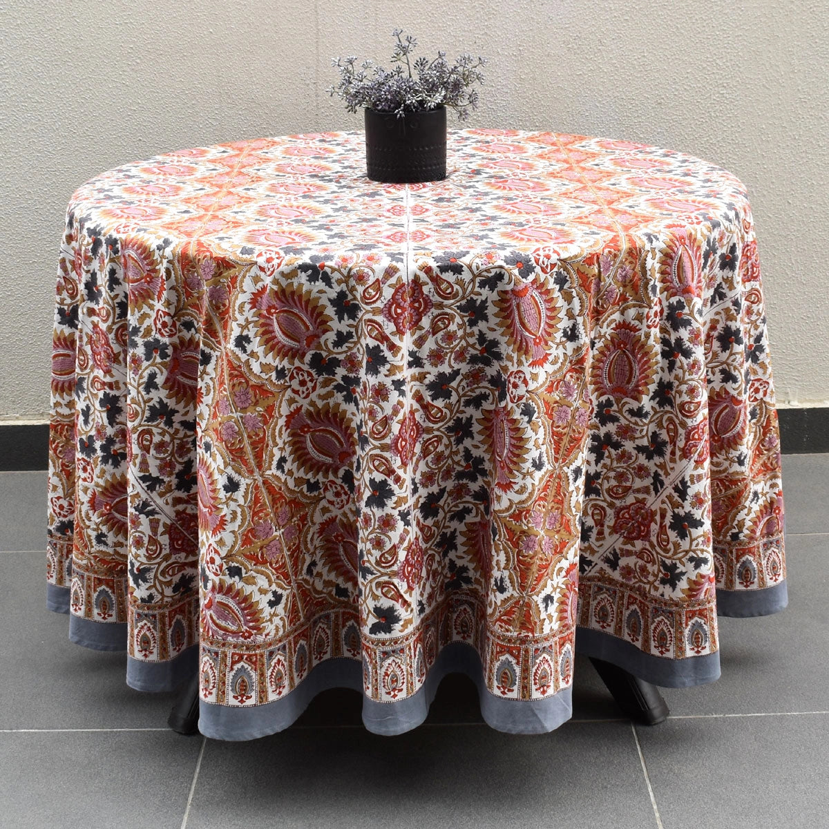 A round table covered with a patterned tablecloth featuring a detailed paisley and floral design in rust red, orange, beige, and gray, with a small black vase of dried stems on top.