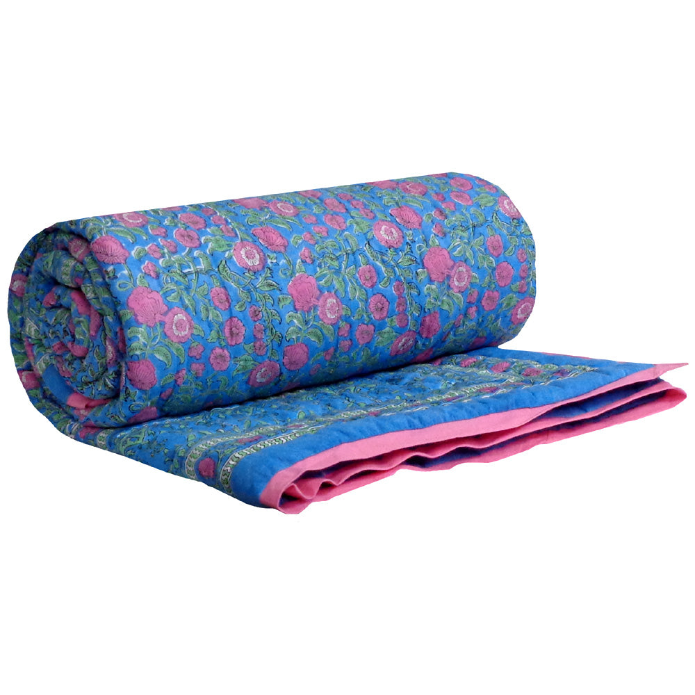 A rolled-up quilted blanket with a vibrant blue and pink floral pattern, shown partially unrolled to display its pink solid inner side on a white background.