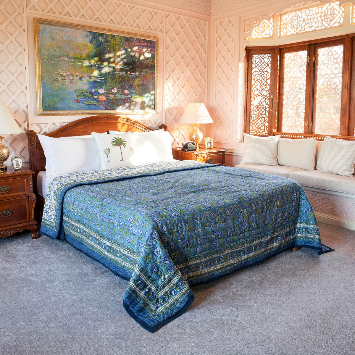 A blue patterned quilted bedspread with matching blue and green pillows is styled on a wooden bed in a decorated bedroom with pink patterned walls, a window with carved wooden screens, and a painting above the headboard.