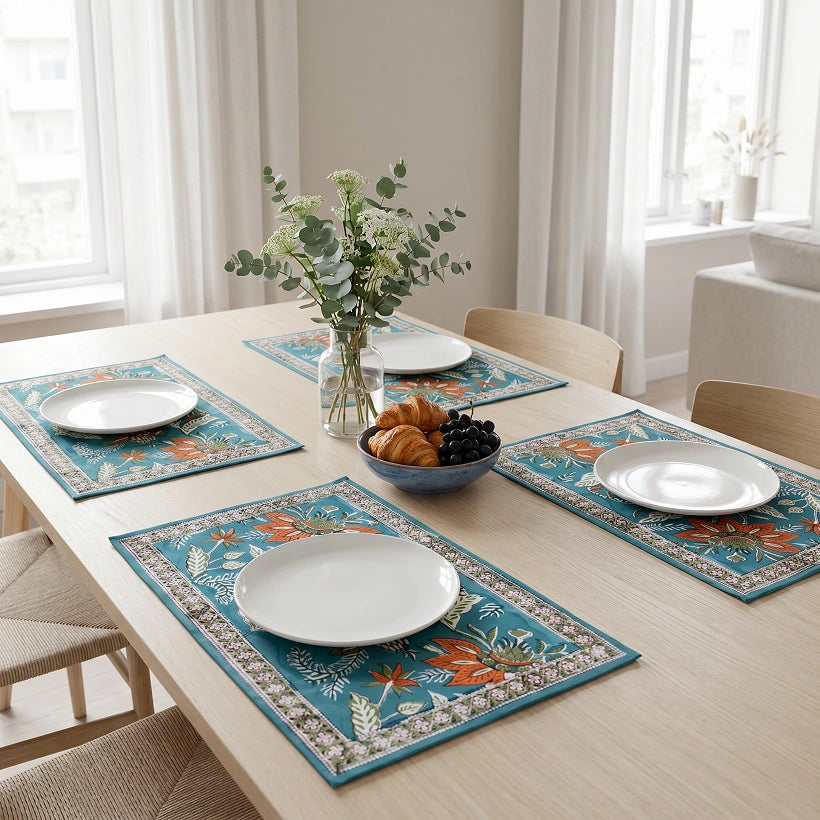 Four rectangular table placemats with a teal blue background and orange, green, and beige floral border design, arranged on a light wood dining table with white plates, a bowl of pastries and grapes, and a vase of greenery in a bright dining room.