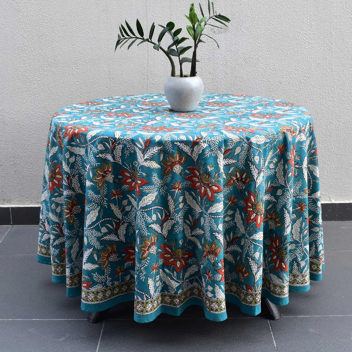 A round table covered with a turquoise blue tablecloth featuring a white and red botanical floral pattern, styled with a white vase of green leaves on top, set against a neutral gray wall and dark tiled floor.