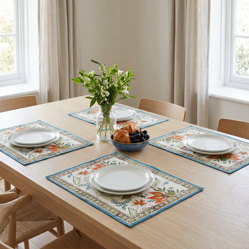 Four rectangular table placemats with a floral and botanical border design in blue, orange, and cream, arranged on a light wood dining table with plates, a vase of flowers, and chairs in a bright room.