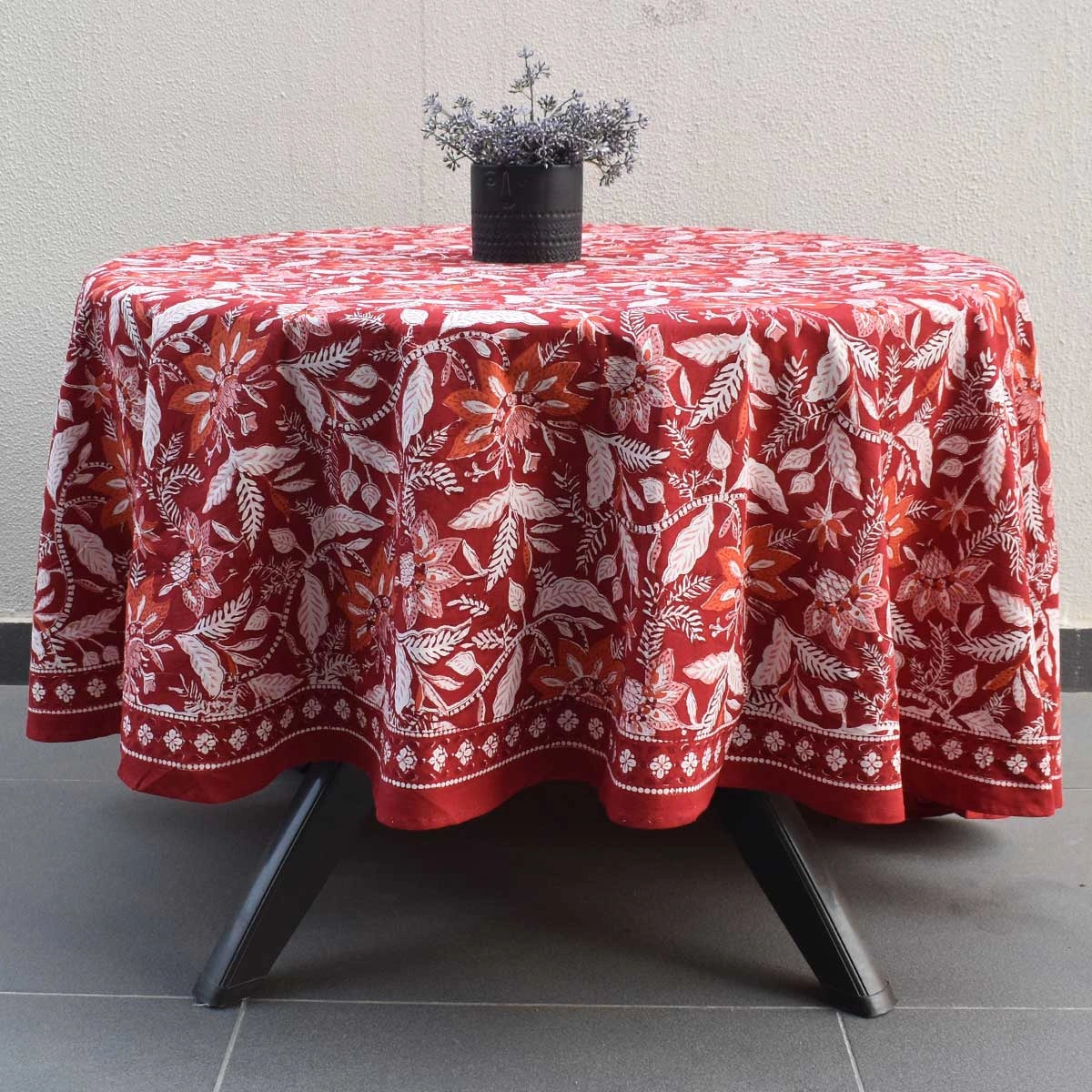 A round table covered with a red tablecloth featuring a white botanical leaf pattern, set on a black metal table with a small black pot of dried flowers on top.
