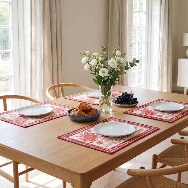 Red and white patterned placemats set on a light wood dining table in a bright, sunlit room with chairs, a vase of white flowers, and a bowl of croissants.