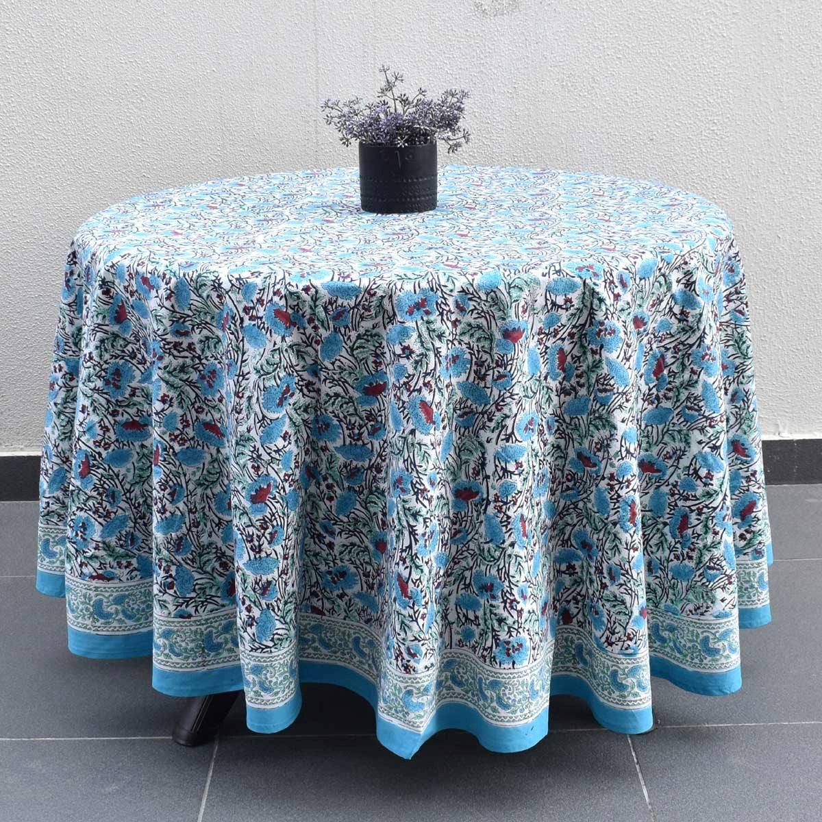 A round table covered with a light blue and white floral patterned tablecloth, styled with a small black vase of dried lavender on top, set against a neutral textured wall and dark tiled floor.