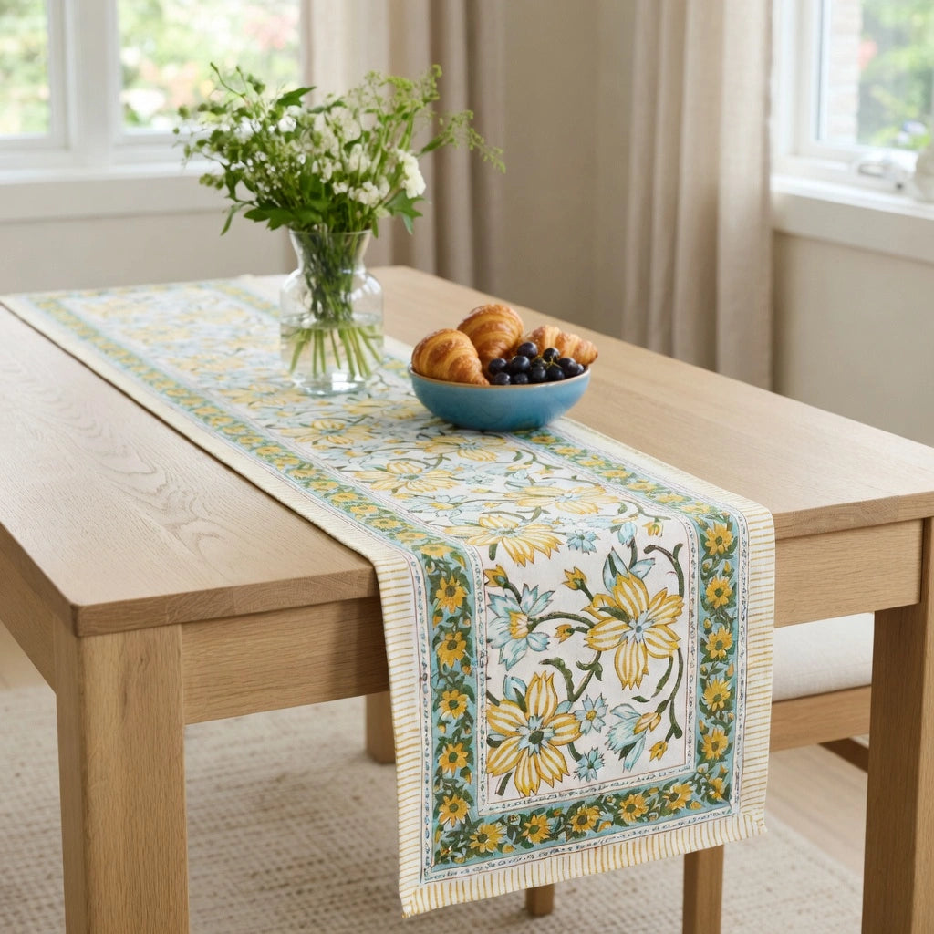 A floral table runner with yellow flowers and green leaves laid across a light wood dining table, styled with a blue bowl of fruit and a vase of white flowers in a bright room.