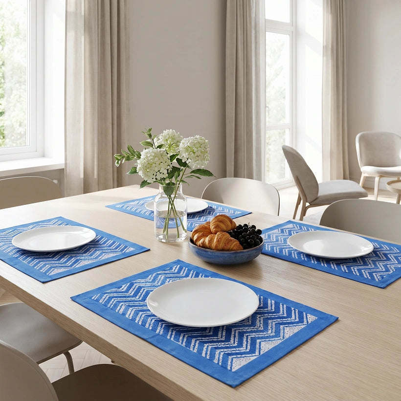 Blue and white patterned woven placemats set on a light wood dining table in a bright, modern room with beige chairs, curtains, and a vase of white flowers.