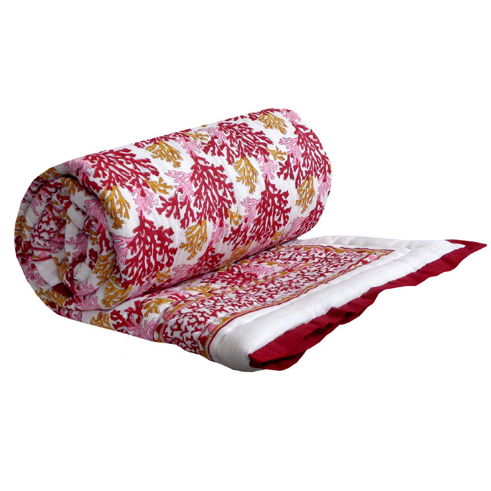 A rolled and partially unrolled quilted blanket featuring a coral-like floral pattern in red, pink, and gold on a white background, with a solid red and white border.