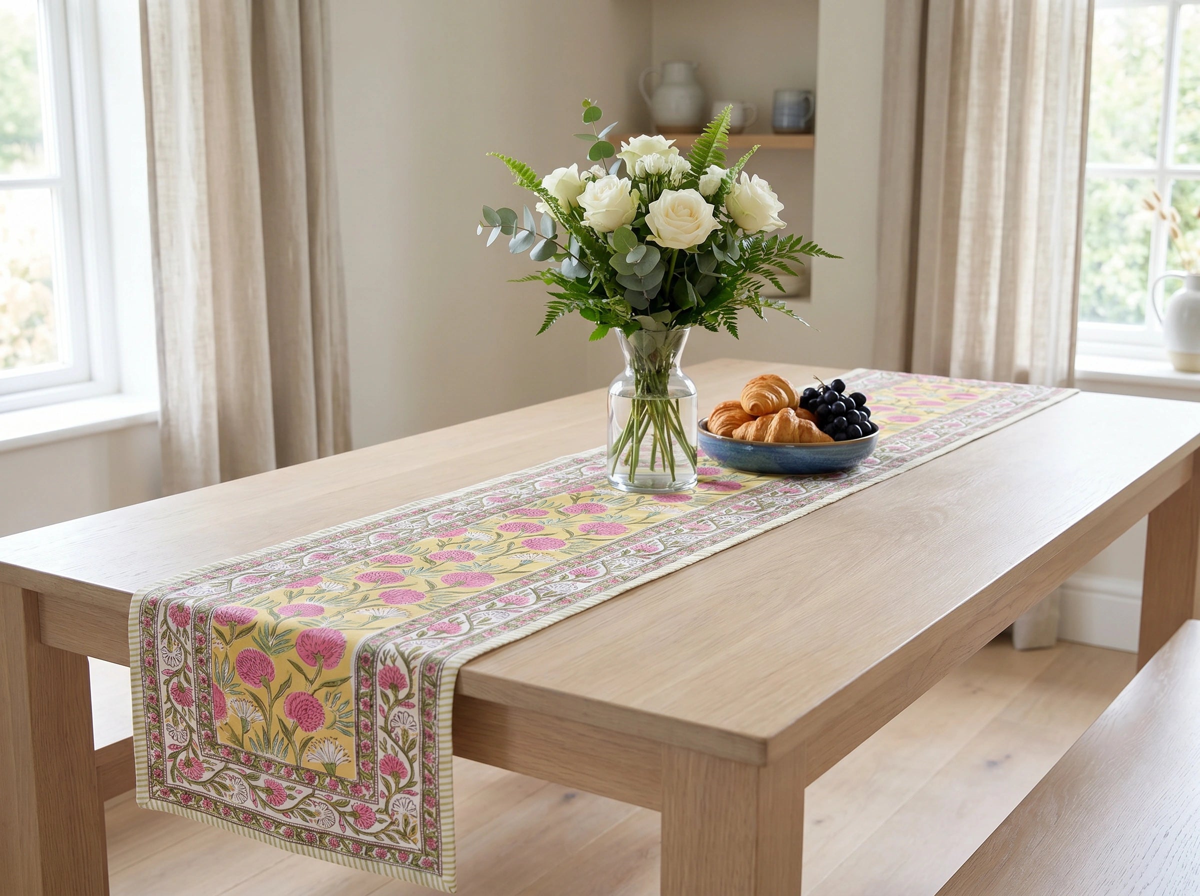 A rectangular table runner with a pink and yellow floral pattern and a decorative border, displayed on a light wood dining table in a bright room with a vase of white roses and a plate of fruit.
