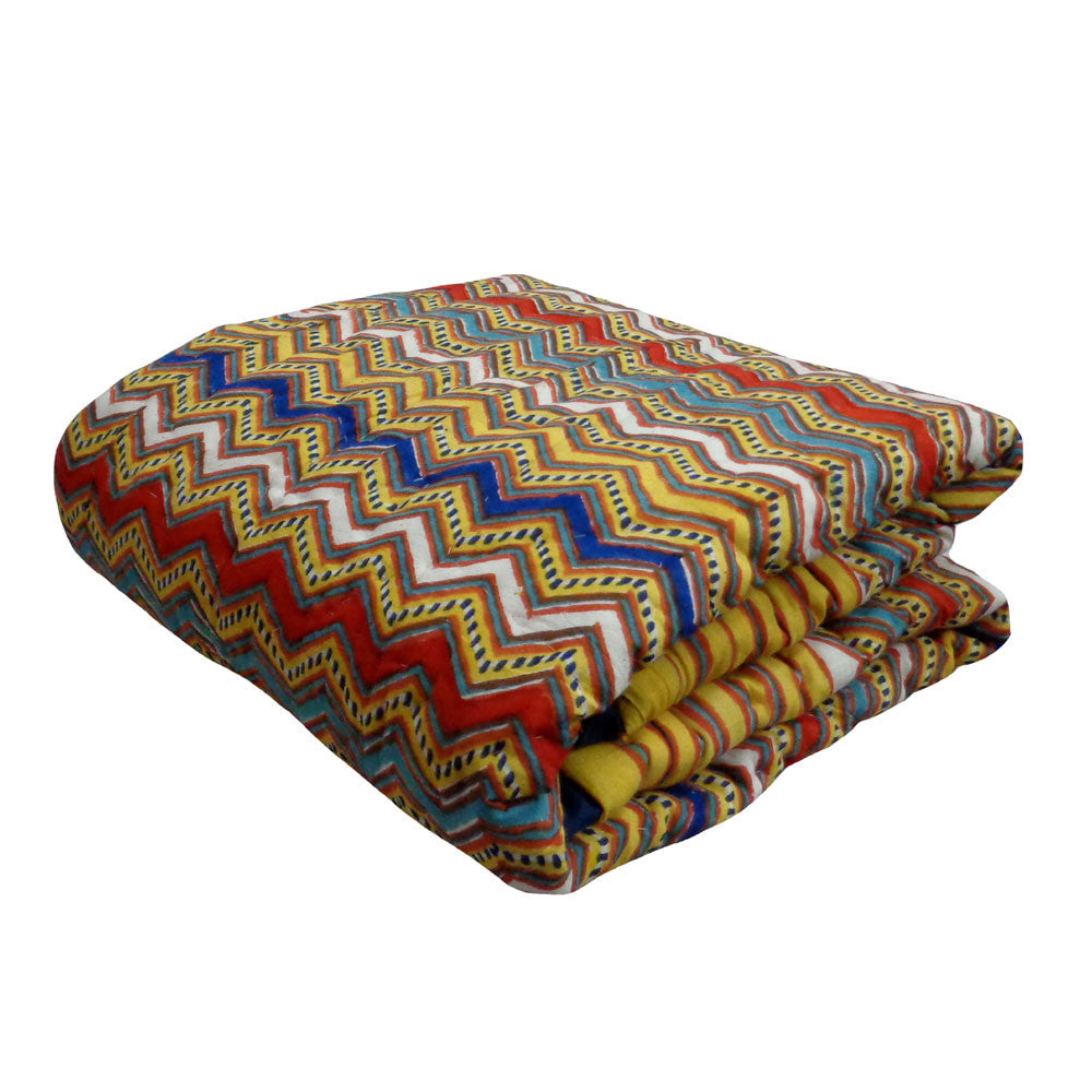 A folded multicolor quilted blanket featuring a vibrant zigzag chevron pattern in yellow, red, blue, white, and black, set against a plain white background.