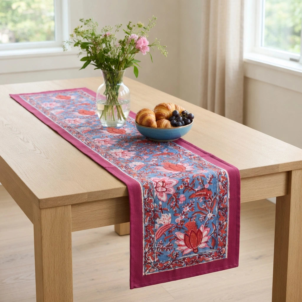 A rectangular table runner with a vibrant blue and red floral pattern and magenta borders, styled on a light wood dining table with a vase of flowers and a bowl of fruit in a bright room.
