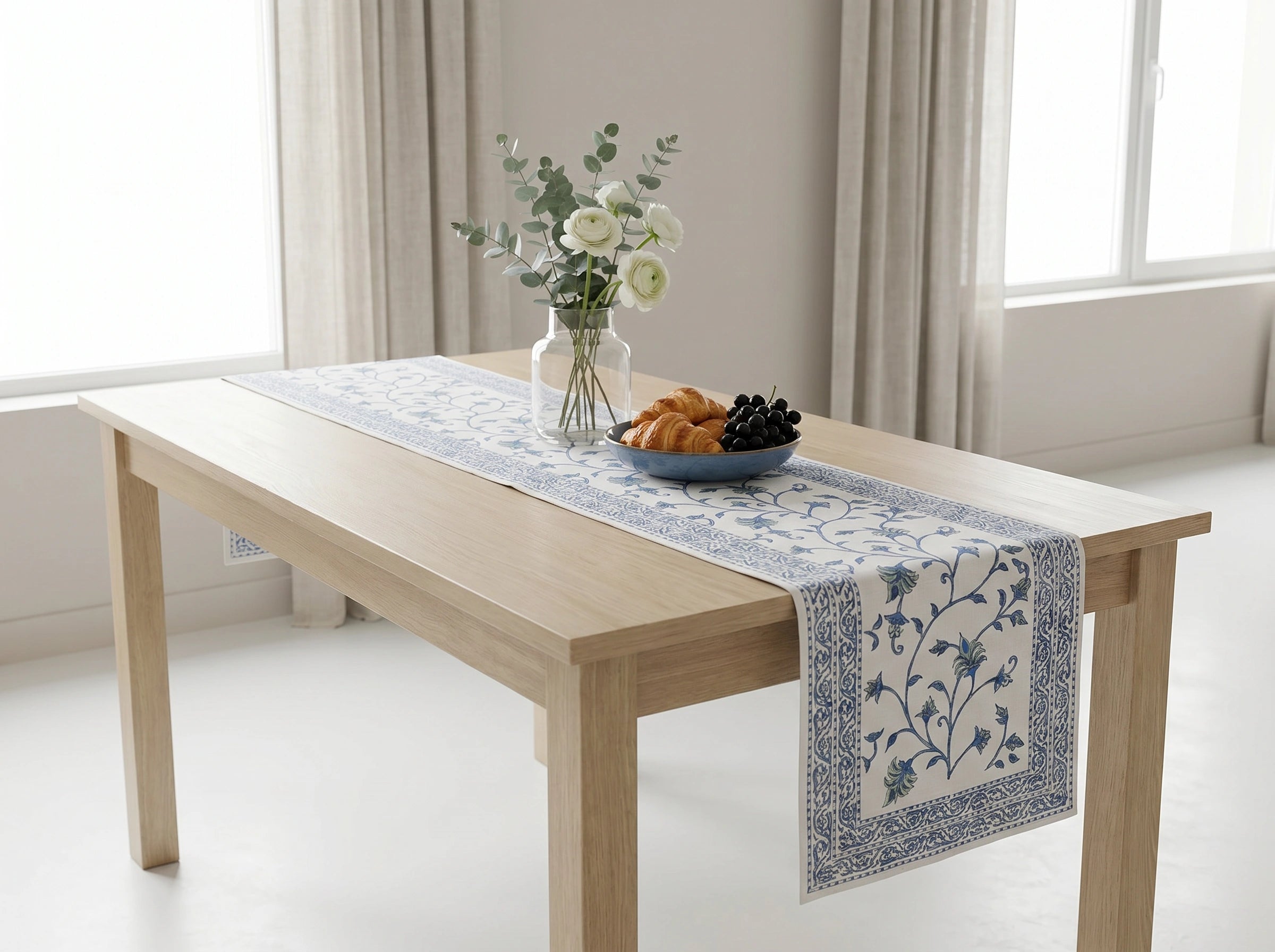 A light wood dining table in a bright room, styled with a blue and white floral table runner, a glass vase with white flowers, and a bowl of fruit.