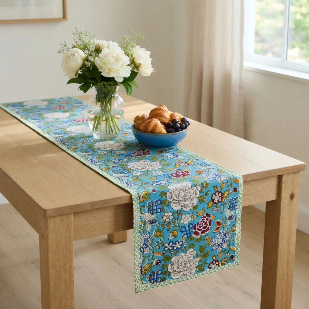 A rectangular embroidered table runner with a floral pattern in shades of blue, green, white, and red, styled on a light wood dining table next to a vase of white flowers and a bowl of pastries.