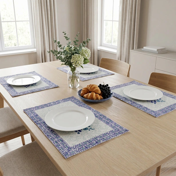 Four rectangular table placemats with blue floral borders and pale blue centers are arranged around a light wood dining table set with white plates, a vase of flowers, and a plate of pastries in a bright dining room.