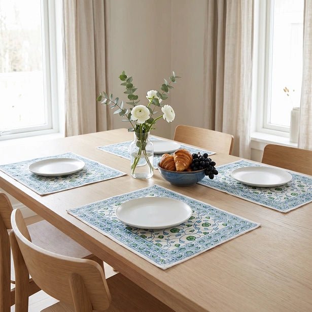 Four rectangular table placemats with a blue and green floral pattern are arranged on a light wood dining table set with white plates, a vase of flowers, and a bowl of pastries in a bright room with beige curtains.