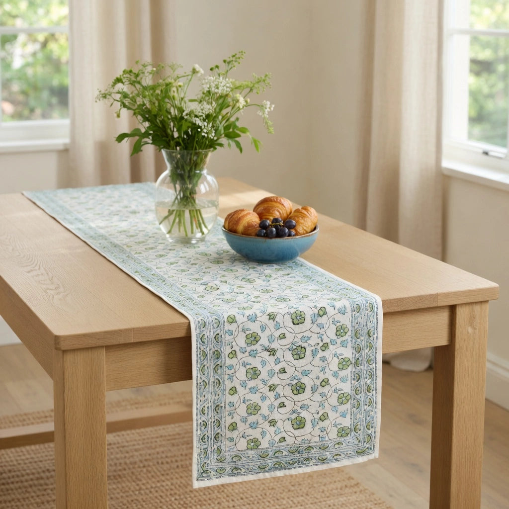 A light blue and white patterned table runner draped over a light wood dining table, styled with a glass vase of greenery and a blue bowl of pastries in a bright room.