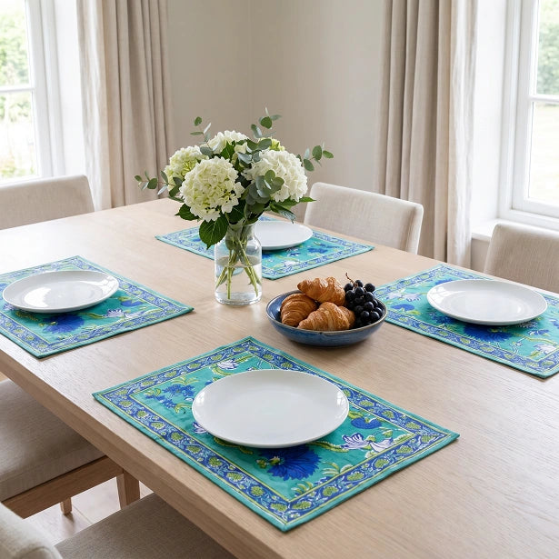 Four matching rectangular placemats with a turquoise and blue floral pattern are arranged on a light wood dining table, each styled with a white plate, a vase of white flowers, and a bowl of pastries in a bright dining room.