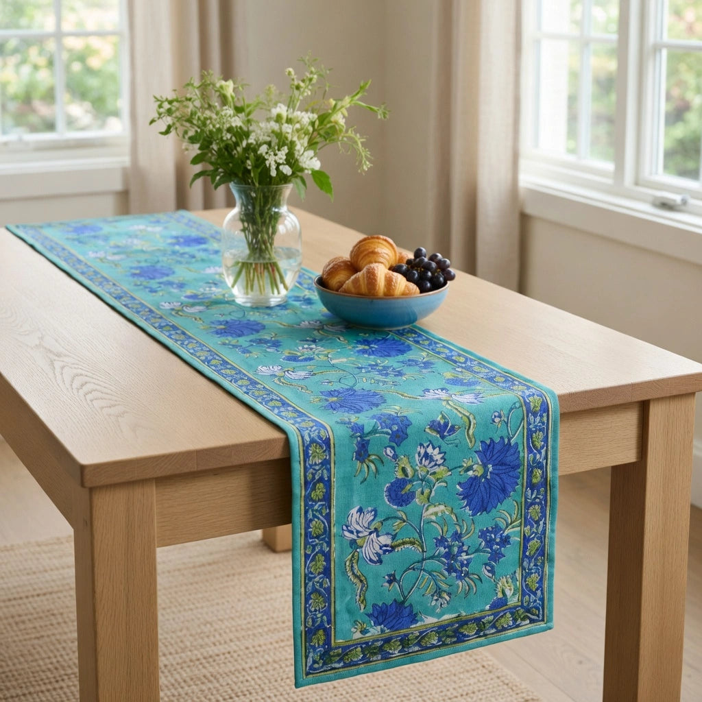 A turquoise and blue floral table runner with a decorative border is draped over a light wood dining table in a bright room. A glass vase with white flowers and a blue bowl of pastries and grapes sit on the table.