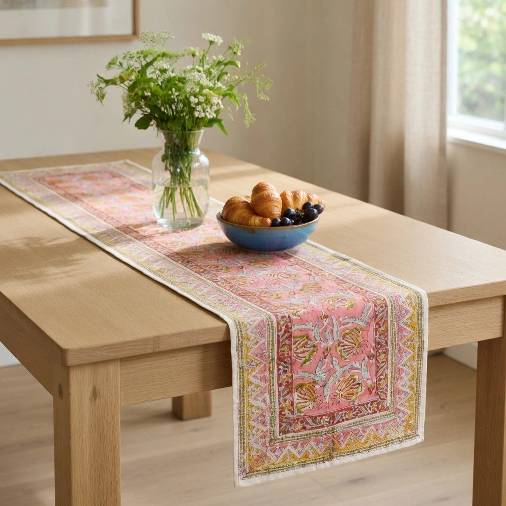 A pink and gold patterned table runner with fringe is draped over a light wood dining table, styled with a glass vase of white flowers and a blue bowl of pastries in a bright room.