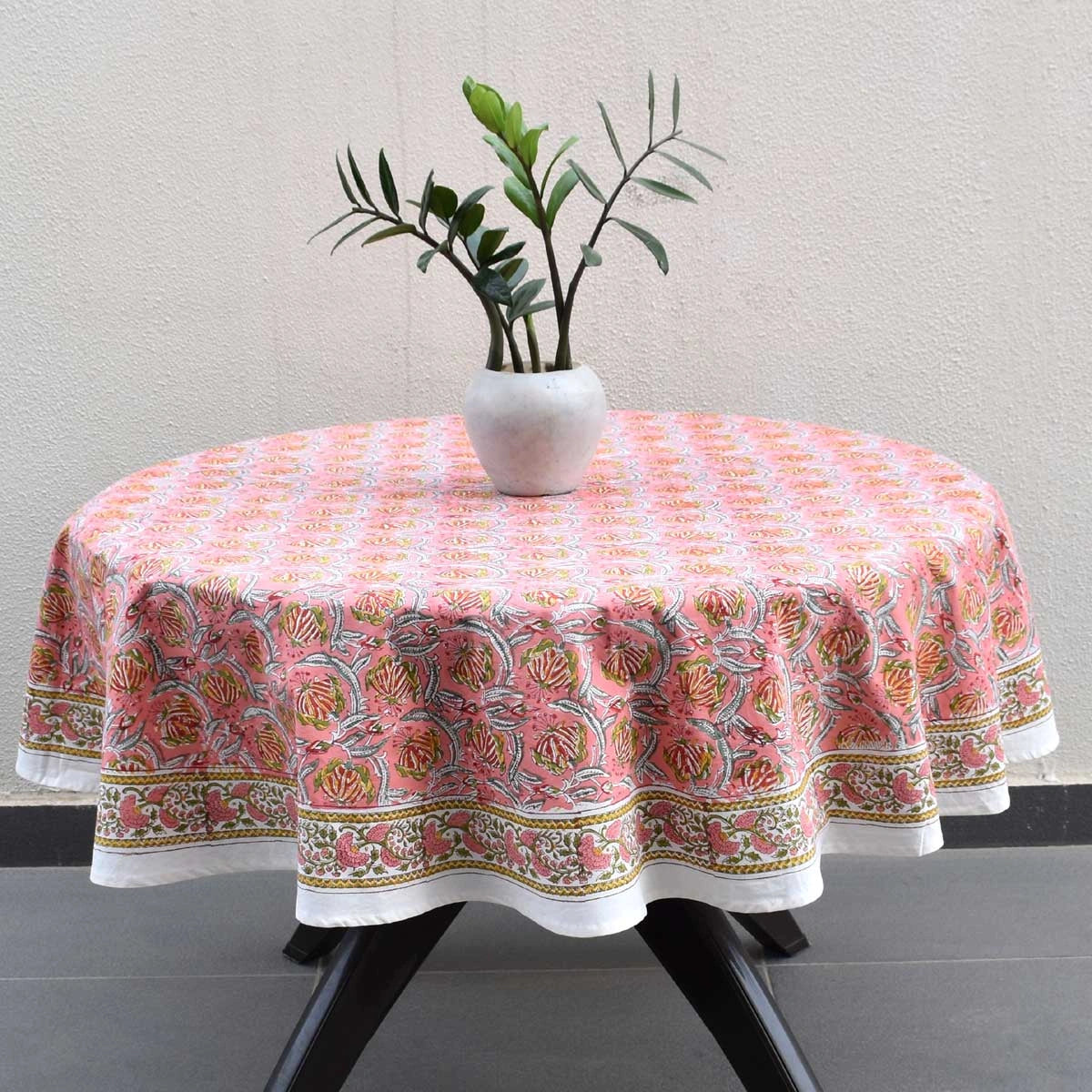 A round table covered with a pink and orange floral patterned tablecloth, styled with a white vase containing green leaves, set against a neutral textured wall.