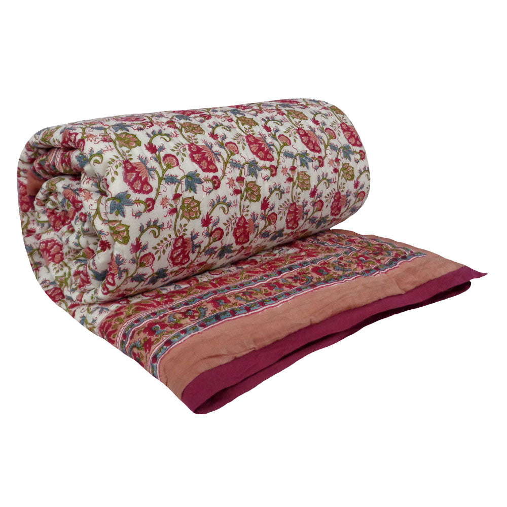 A rolled-out quilted blanket featuring a traditional floral block print pattern in red, green, and blue on an off-white background, with a solid dusty pink border and a deep magenta underside.