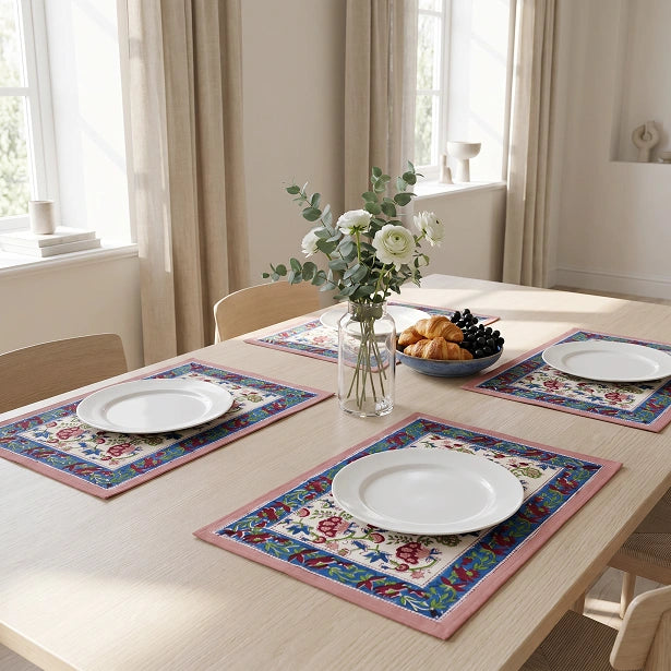 Three rectangular table placemats with a colorful floral and geometric border design are arranged on a light wood dining table, each styled with a white plate, a glass vase of flowers, and a bowl of pastries in a bright room.