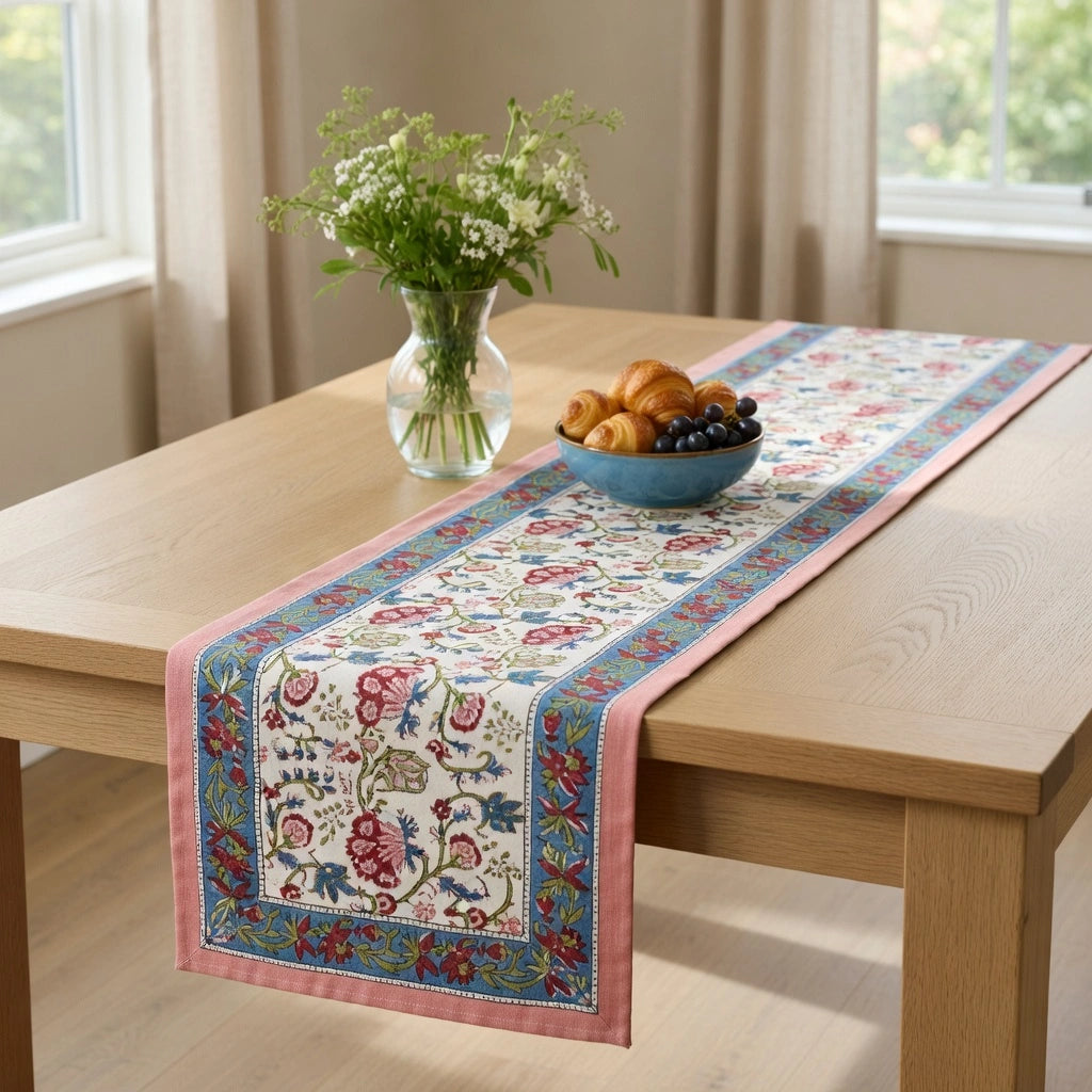 A rectangular table runner with a pink border and a detailed floral pattern in red, blue, and green on a cream background, styled on a light wood dining table with a vase of flowers and a bowl of pastries.
