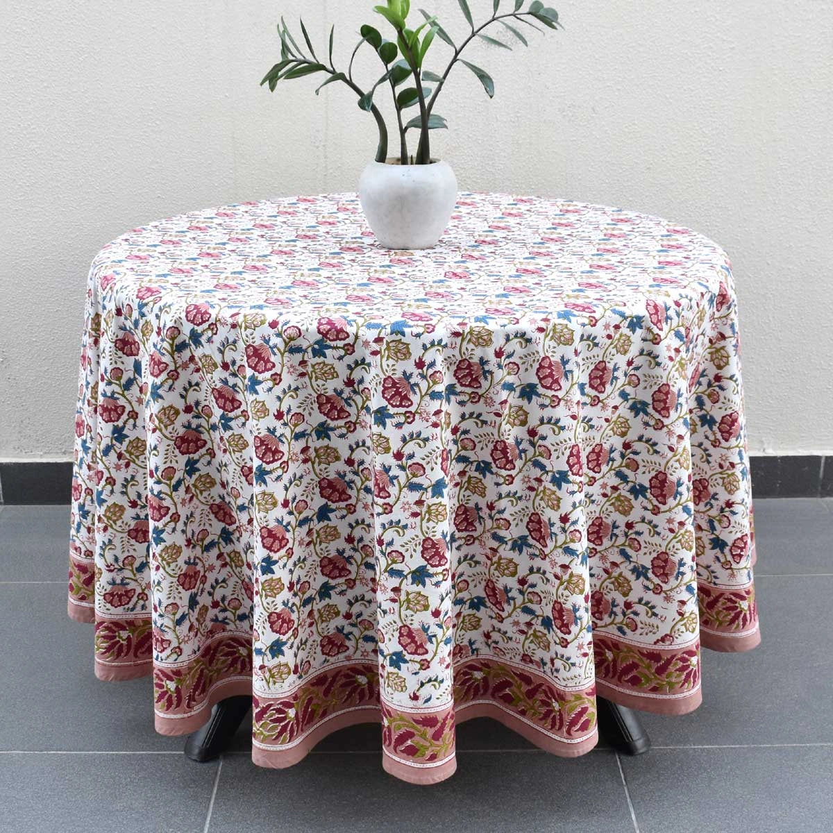 A round table covered with a white tablecloth featuring a dense pink, red, and blue floral pattern and a decorative border, styled with a white vase of green leaves on top.