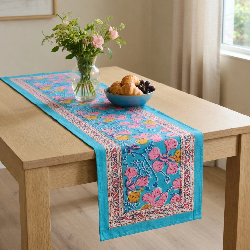 A turquoise blue table runner with pink floral and yellow butterfly patterns, styled on a light wood dining table with a vase of flowers and a bowl of pastries.