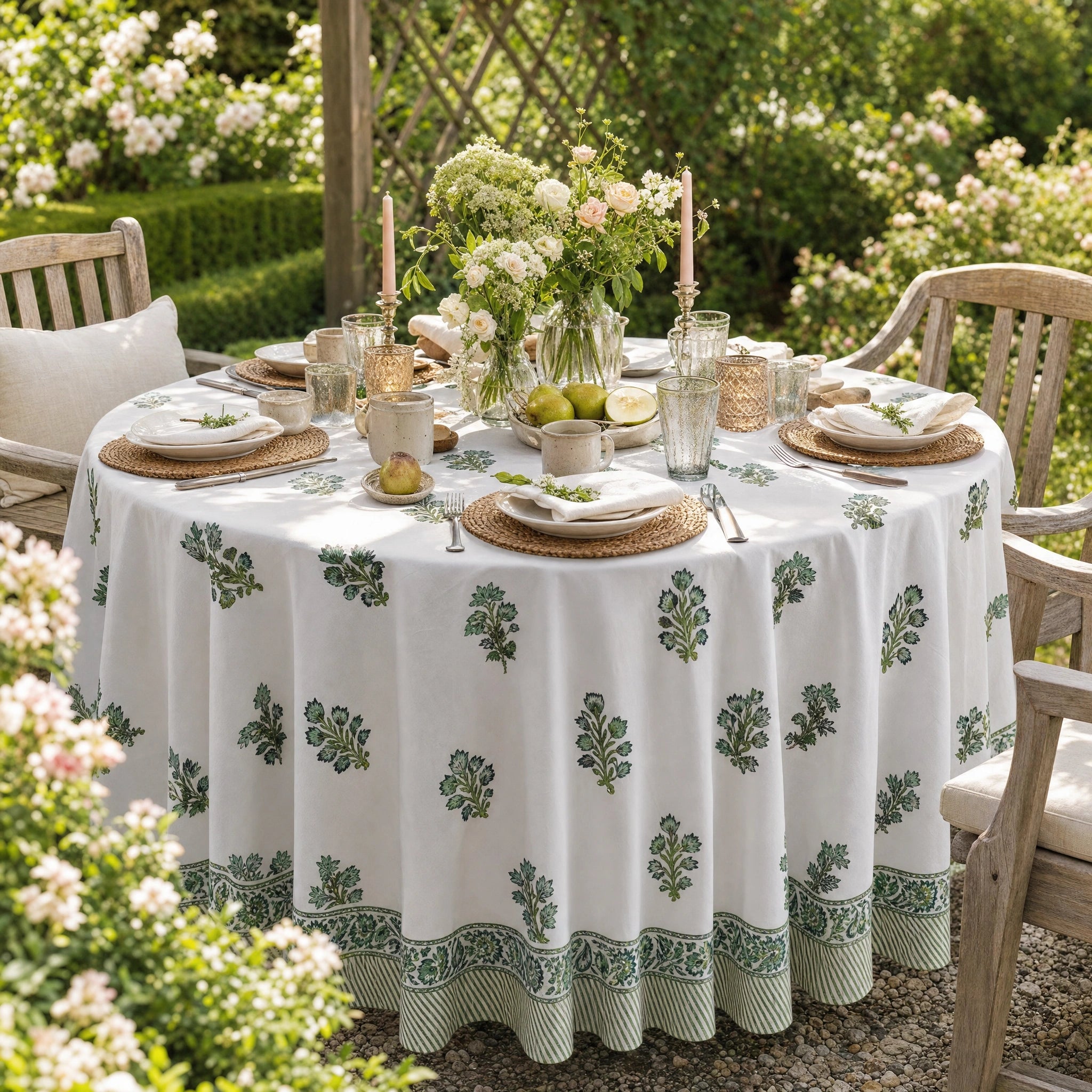 A round outdoor dining table covered with a white tablecloth printed with green botanical motifs and a striped border, set with plates, glasses, candles, and a floral centerpiece in a garden patio setting.