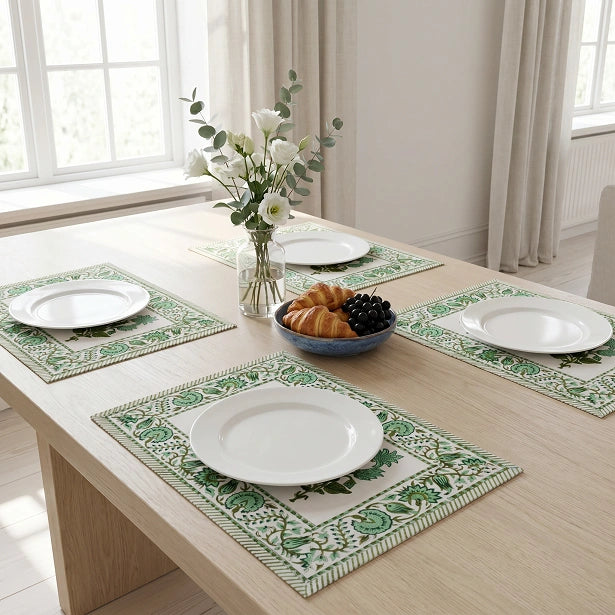 Four rectangular table placemats with green botanical patterns and striped borders are arranged on a light wood dining table. Each placemat holds a white plate, and a small bowl of fruit and a vase of white flowers sit in the center.