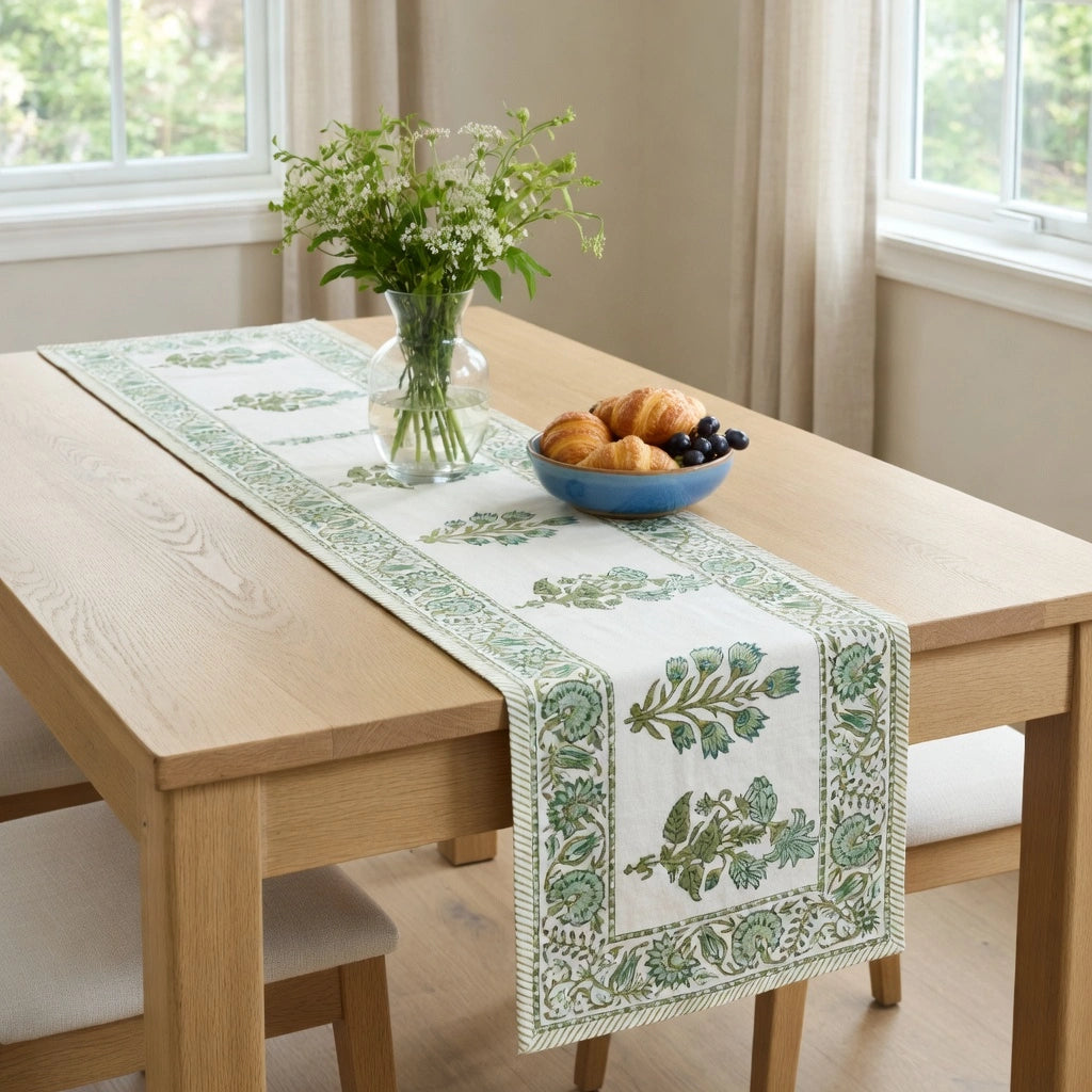 A rectangular table runner with a green botanical border and central leaf pattern, styled on a light wood dining table with a vase of flowers and a bowl of pastries.