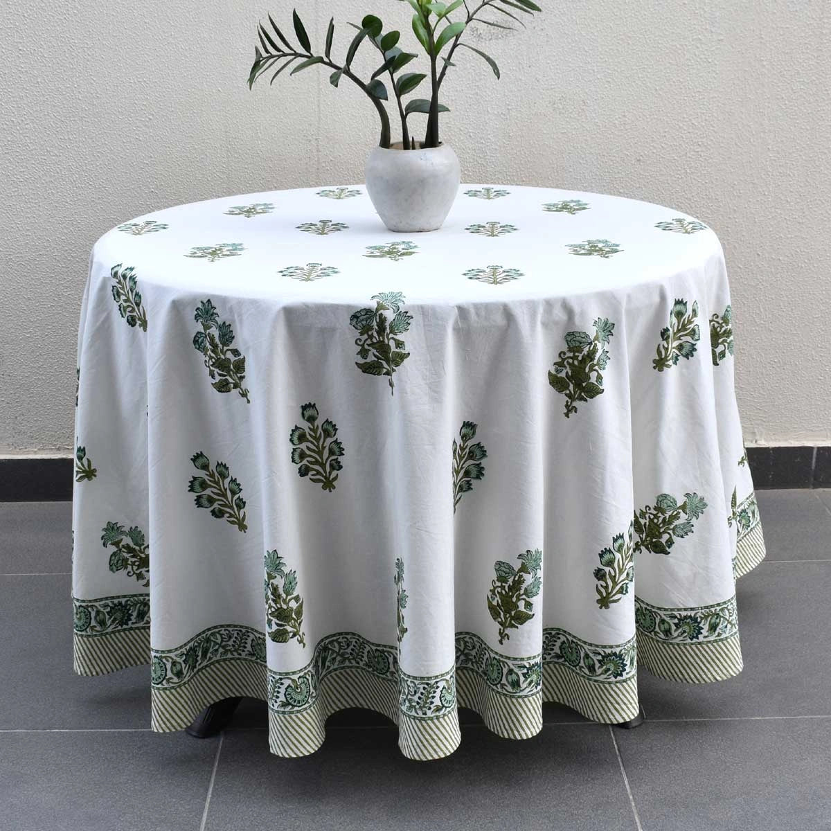 A round white tablecloth with a green botanical block print pattern and a decorative border, draped over a round table with a small white vase of green leaves on top, set against a neutral wall and dark tiled floor.