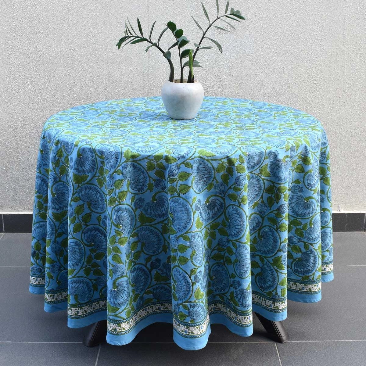 A round table covered with a blue and green floral patterned tablecloth, styled with a small white vase containing green leaves, set against a neutral textured wall and dark tiled floor.