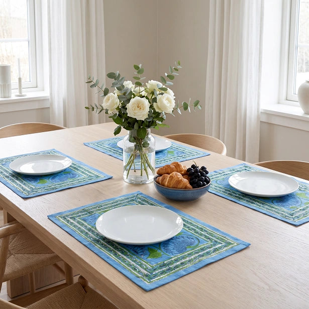 A dining table set with four blue patterned placemats, white plates, a glass vase of white flowers, and a bowl of pastries in a bright, modern dining room.