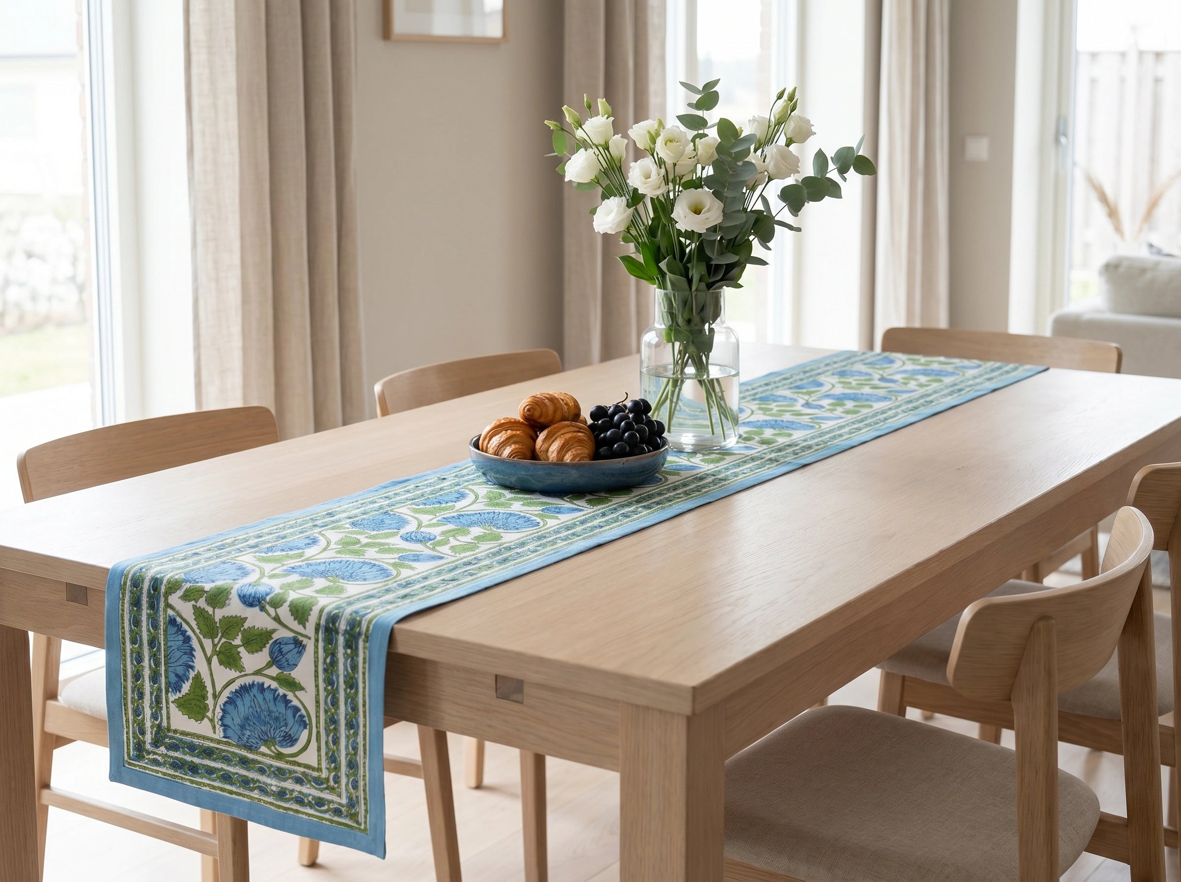 A light blue and green floral table runner with a decorative border, styled on a light wood dining table with a vase of white flowers and a bowl of fruit in a bright, modern dining room.