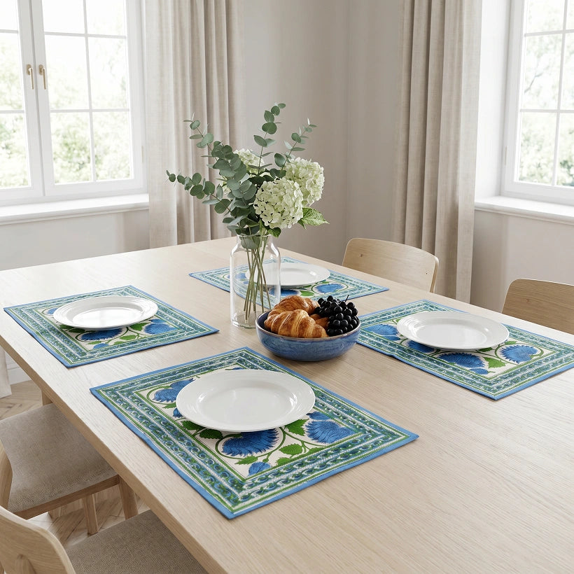 Three decorative placemats with blue and green floral patterns are arranged on a light wood dining table, each styled with a white plate, a bowl of pastries and grapes, and a vase of flowers in a bright room with windows and curtains.