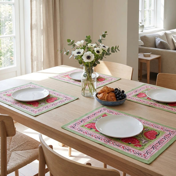 Four rectangular table placemats with a pink floral and green border design are arranged on a light wood dining table set with white plates, a vase of white flowers, and a bowl of pastries in a bright, modern dining room.