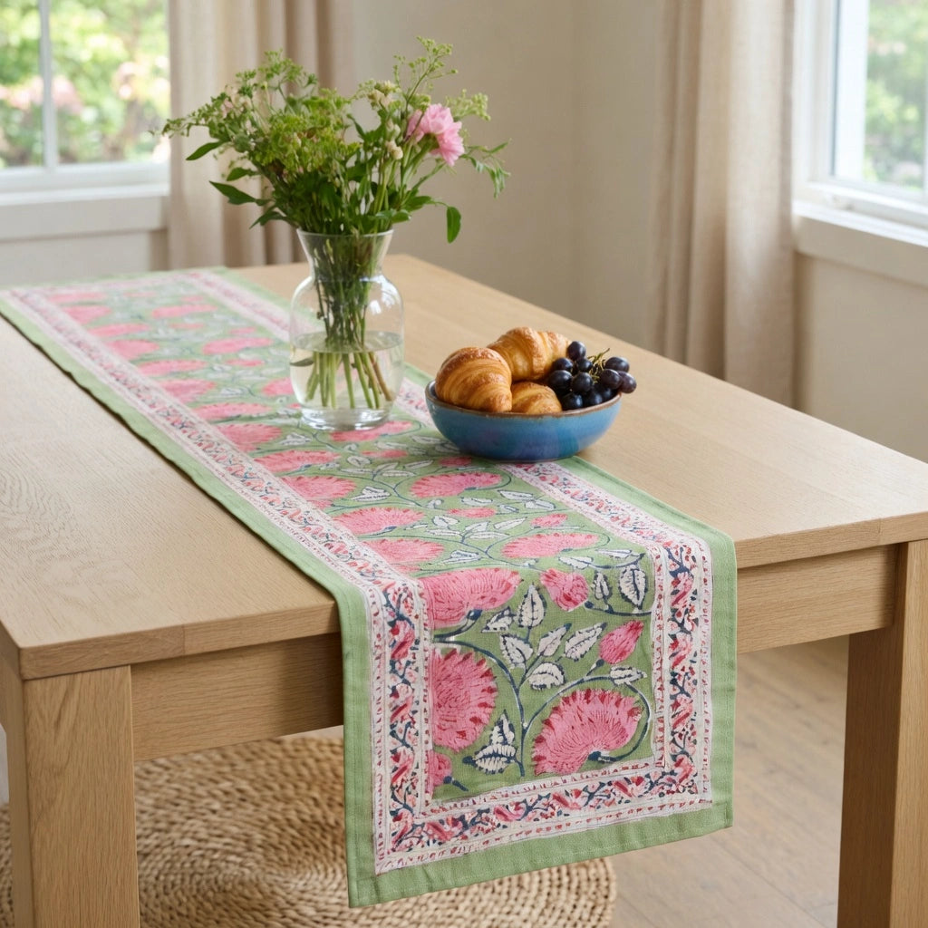 A light green table runner with pink floral and blue border patterns is styled on a light wood dining table. The table also features a glass vase with green stems and a blue bowl of pastries and grapes, set in a bright room with beige curtains and windows.