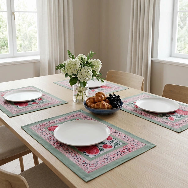 Three rectangular table placemats with a floral and ornamental border design in sage green, pink, and red are arranged on a light wood dining table. Each placemat holds a white plate, and the setting includes a vase of white flowers, a bowl of pastries, and wooden chairs in a bright dining room.