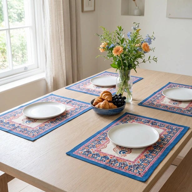 Four rectangular table placemats with a blue and cream traditional pattern are arranged on a light wood dining table. Each placemat holds a white plate, with a central vase of flowers and a bowl of pastries on the table.