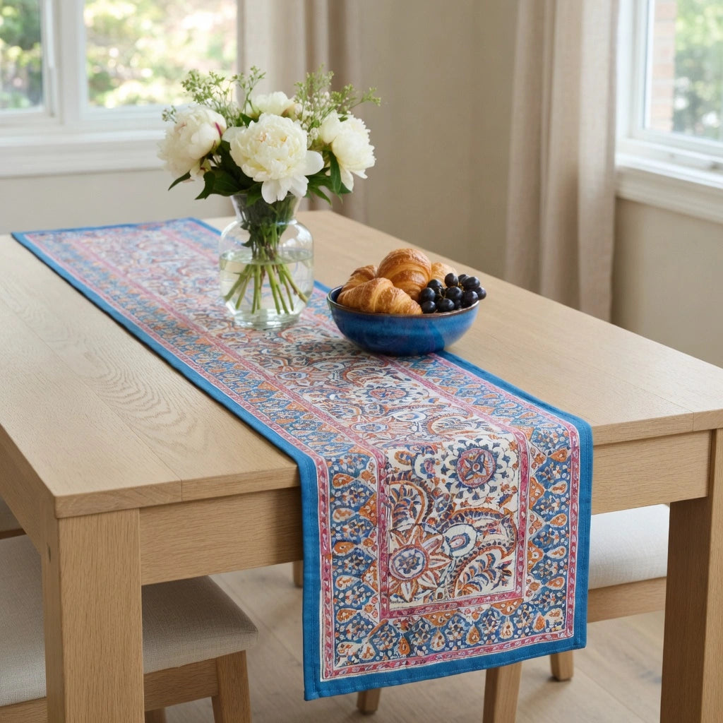 A rectangular table runner with a detailed blue, red, and beige paisley pattern is styled on a light wood dining table. The table runner is decorated with a glass vase of white flowers and a blue bowl of pastries, set in a bright room with beige walls and curtains.
